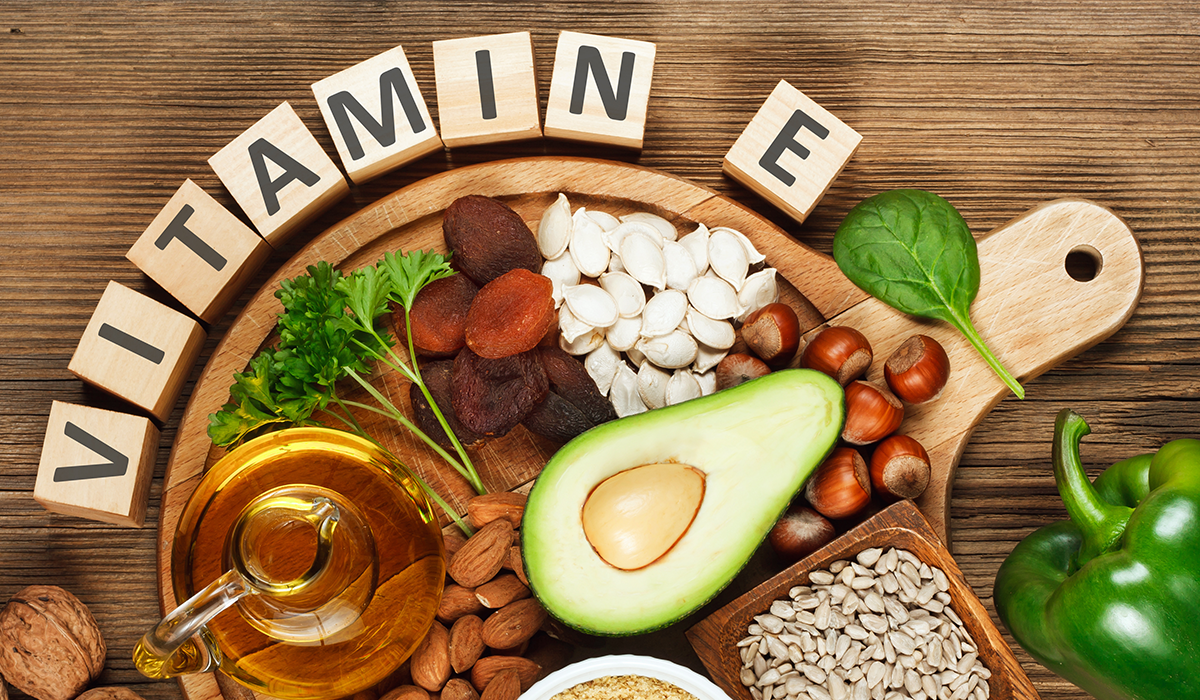 En bild med ordet Vitamin E i en halvmåne runt en skärbräda med massa livsmedel som innehåller en hög halt av Vitamin E.